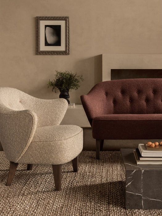 An Ingeborg Sofa in rust and Ingeborg Lounge Chair in cream styled in a living room on a jute rug beside a marble table.