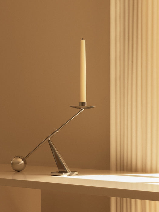 The small, sculptural Interconnect Candle Holder in polished steel holds a cream candle on a shelf in warm, soft light.