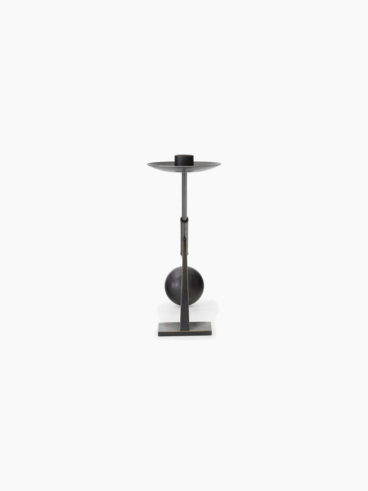 A tall, sculptural candle holder in dark, bronzed metal with a rectangular base, a central sphere, and a slender stem.