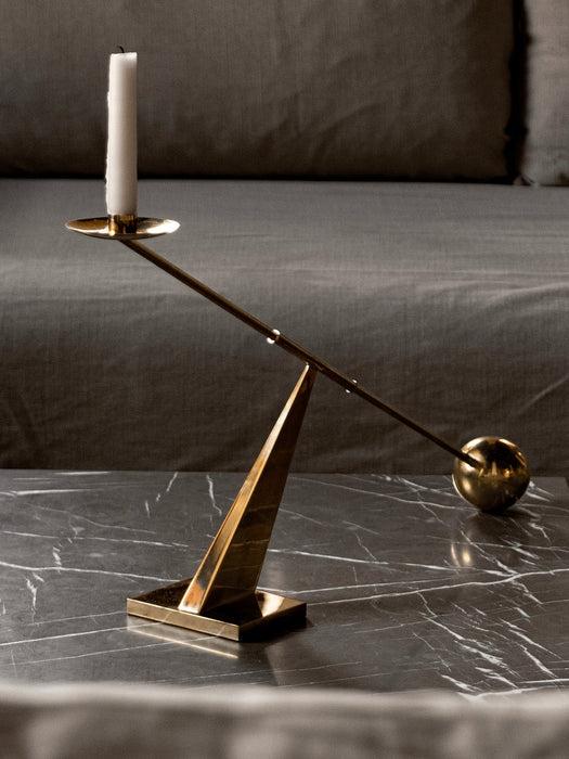 The brass Interconnect Candle Holder, with a white candle, rests on a black marble surface, resembling a balance scale.