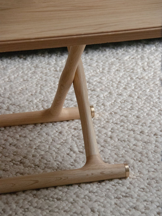 Close-up of the Jäger Lounge Table's natural oak legs with brass fittings, showing the criss-cross design on a cream rug.