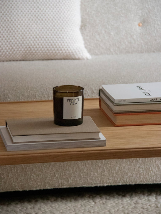 A close-up of a wooden Jäger Lounge Table, styled with a Private View scented candle and books next to a beige sofa.