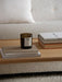 A close-up of a wooden Jäger Lounge Table, styled with a Private View scented candle and books next to a beige sofa.