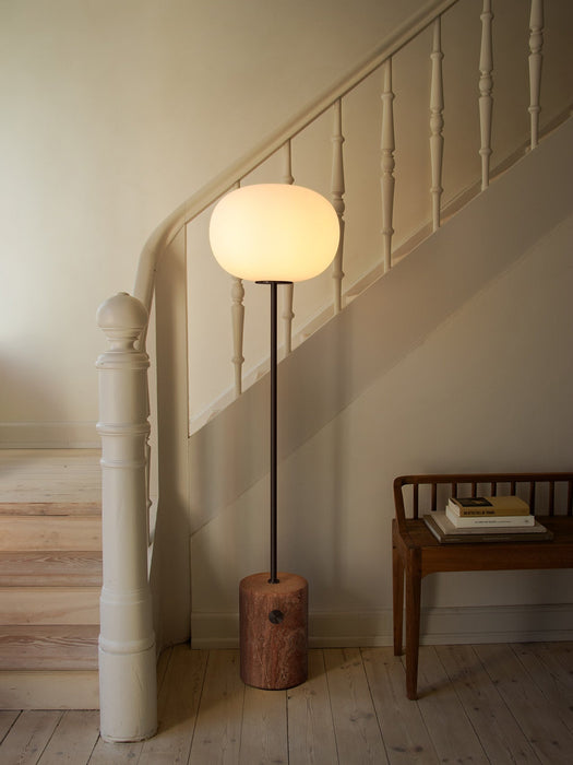 The JWDA Floor Lamp with a red travertine base and glowing opal shade, standing on a wooden floor next to a white staircase.