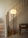 The JWDA Floor Lamp with a red travertine base and glowing opal shade, standing on a wooden floor next to a white staircase.
