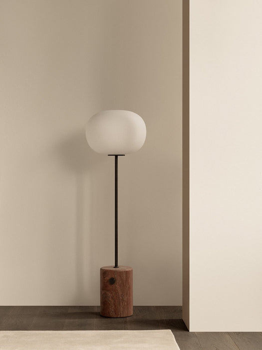 The JWDA Floor Lamp with a red travertine base and opal glass shade, standing on a dark wood floor against a beige wall.