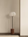 The JWDA Floor Lamp with a red travertine base and opal glass shade, standing on a dark wood floor against a beige wall.