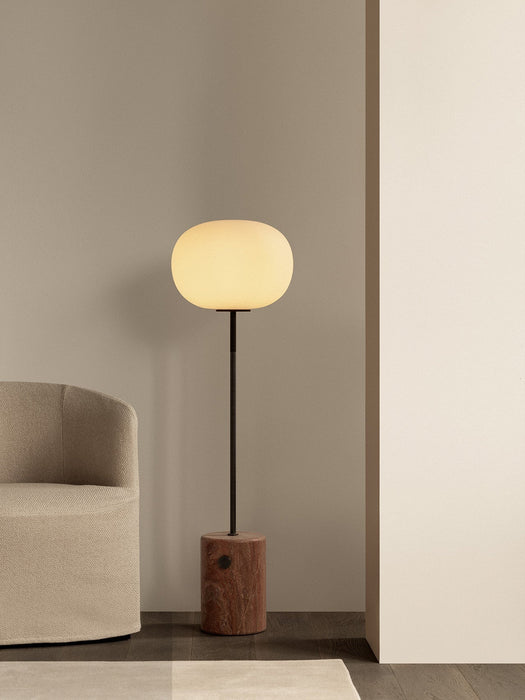 The JWDA Floor Lamp in Red Travertine stands lit in a minimalist room next to a textured beige Tearoom Lounge Chair.