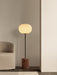 The JWDA Floor Lamp in Red Travertine stands lit in a minimalist room next to a textured beige Tearoom Lounge Chair.