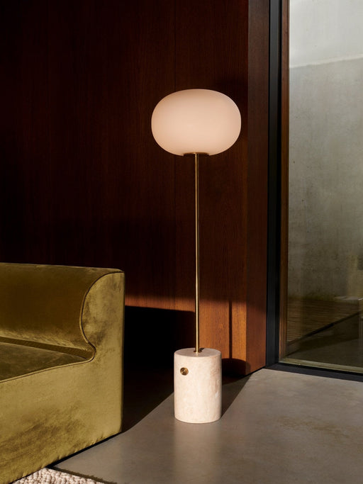 The JWDA Floor Lamp, with travertine base and glowing glass shade, in a sunlit corner beside an olive-green velvet Eave Sofa.