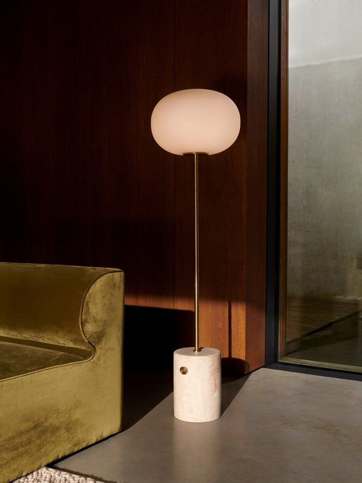 The JWDA Floor Lamp, with travertine base and glowing glass shade, in a sunlit corner beside an olive-green velvet Eave Sofa.
