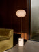 The JWDA Floor Lamp, with travertine base and glowing glass shade, in a sunlit corner beside an olive-green velvet Eave Sofa.