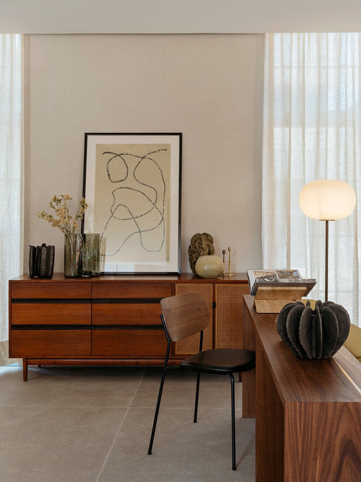 A warmly lit JWDA Floor Lamp in a living room with a wood sideboard, framed abstract art, a modern chair, and sheer curtains.