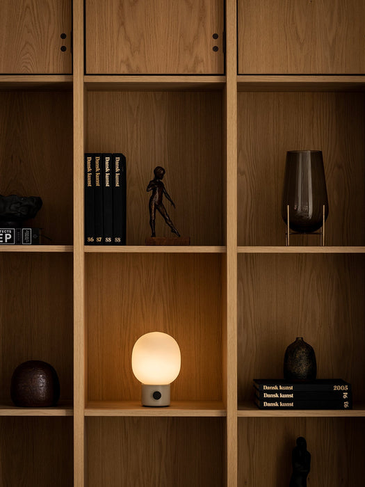 The JWDA Table Lamp, Portable, glowing warmly on a wooden bookshelf, styled with books, sculptures, and a smoked glass vase.