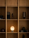The JWDA Table Lamp, Portable, glowing warmly on a wooden bookshelf, styled with books, sculptures, and a smoked glass vase.