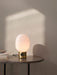 The JWDA Table Lamp in polished brass with an illuminated opal glass shade, on a dark grey table next to a stack of books.