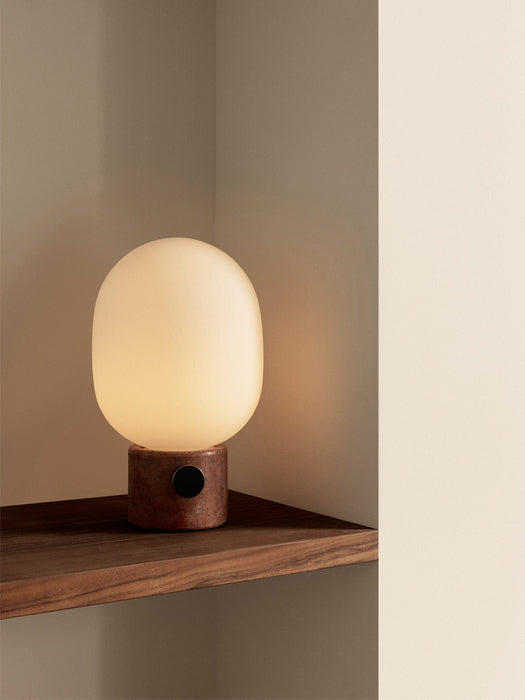 The JWDA Table Lamp, Large, with a Red Travertine base and glowing opal shade, in the corner of a dark wooden shelf.