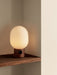 The JWDA Table Lamp, Large, with a Red Travertine base and glowing opal shade, in the corner of a dark wooden shelf.