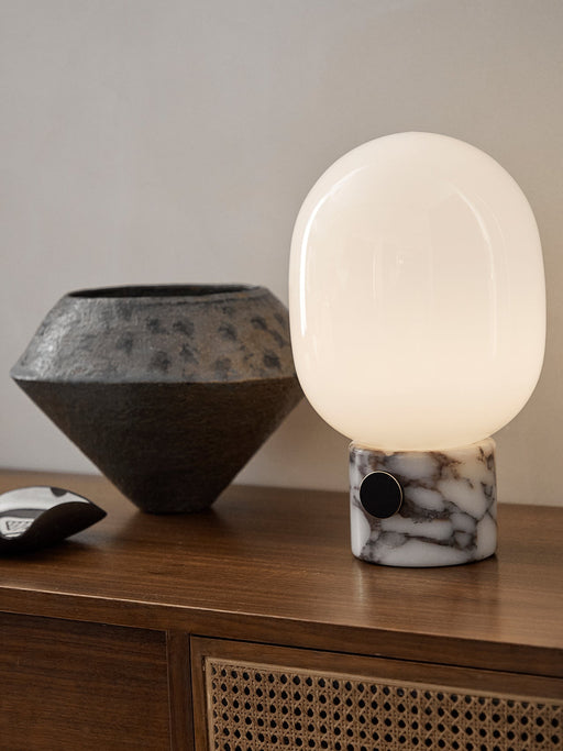 The JWDA Table Lamp, illuminated, with its Calacatta Viola marble base and opal glass shade, sits on a wooden sideboard.