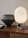 The JWDA Table Lamp, illuminated, with its Calacatta Viola marble base and opal glass shade, sits on a wooden sideboard.