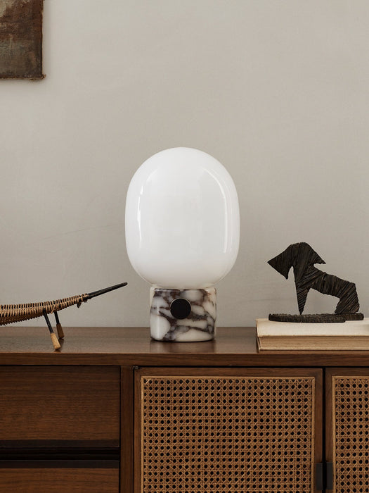 The JWDA Table Lamp in Calacatta Viola marble sits on a dark wood cabinet, styled with a book and abstract sculptures.