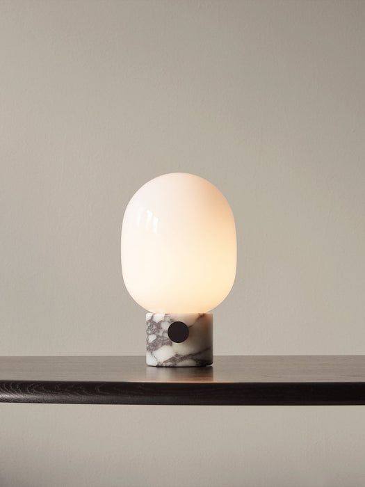 A lit JWDA Table Lamp with an opal glass shade and a veined marble base, standing on a dark wooden shelf against a grey wall.