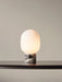 A lit JWDA Table Lamp with an opal glass shade and a veined marble base, standing on a dark wooden shelf against a grey wall.