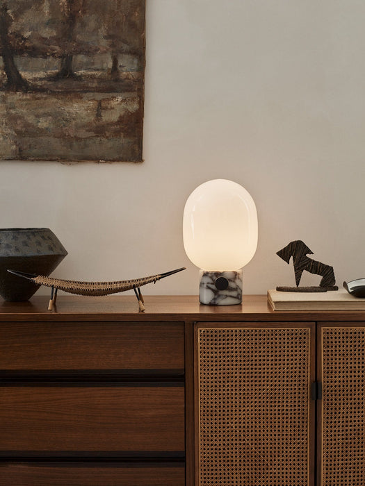 The JWDA Table Lamp in Calacatta Viola marble, lit on a wood sideboard with rattan doors, styled with art and sculptures.