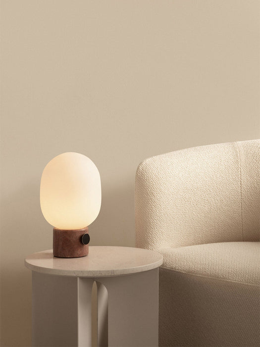 The lit JWDA Table Lamp with a red travertine base sits on an Androgyne Side Table next to a cream Tearoom Lounge Chair.
