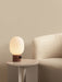 The lit JWDA Table Lamp with a red travertine base sits on an Androgyne Side Table next to a cream Tearoom Lounge Chair.