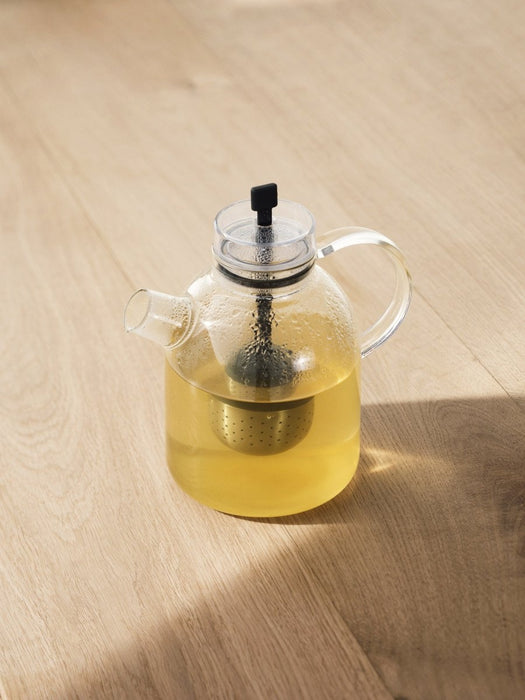 A clear glass Kettle Teapot filled with pale yellow tea, with a metal infuser inside, sitting on a light wood surface.