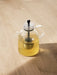 A clear glass Kettle Teapot filled with pale yellow tea, with a metal infuser inside, sitting on a light wood surface.