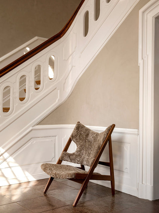 The Knitting Lounge Chair in sheepskin, with its dark wooden frame, sits on a tiled floor by a grand, white staircase.