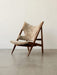 Side view of the Knitting Lounge Chair with a dark stained wood frame and light brown sheepskin upholstery on a grey floor.