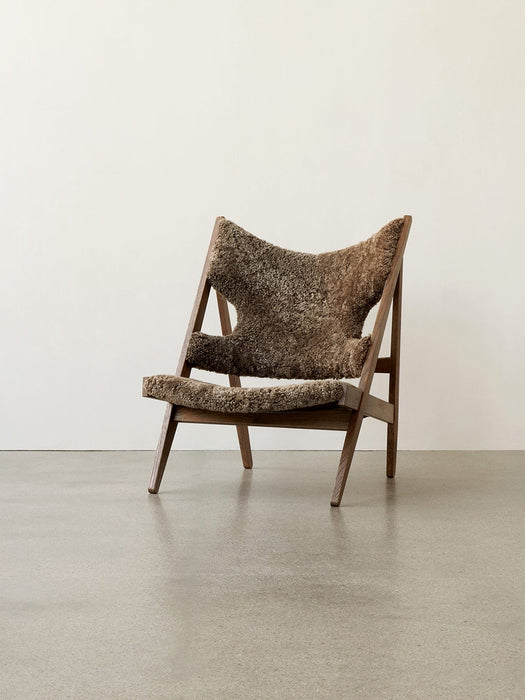 A side view of the Knitting Lounge Chair, upholstered in thick brown sheepskin with a dark wooden frame, on a grey floor.