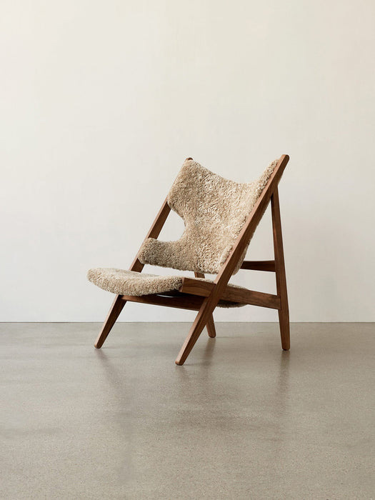 A Knitting Lounge Chair in light Sheepskin with a dark wood frame, viewed from the side on a grey floor against a white wall.