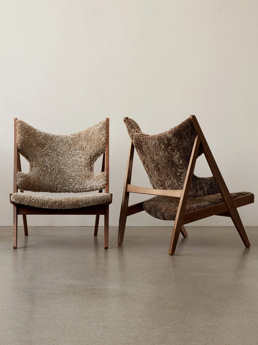 Two Knitting Lounge Chairs in sheepskin with wood frames, one light brown & one dark brown, against a plain off-white wall.