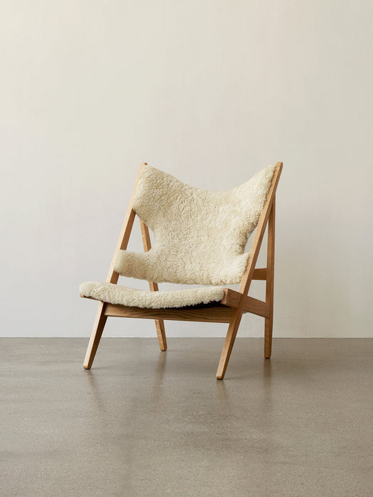 A side-angle view of the Knitting Lounge Chair in natural oak with off-white sheepskin upholstery, against a white wall.