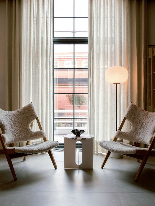Two Knitting Lounge Chairs in Sheepskin flank an Androgyne Side Table in a living room with a tall window and a JWDA Lamp.