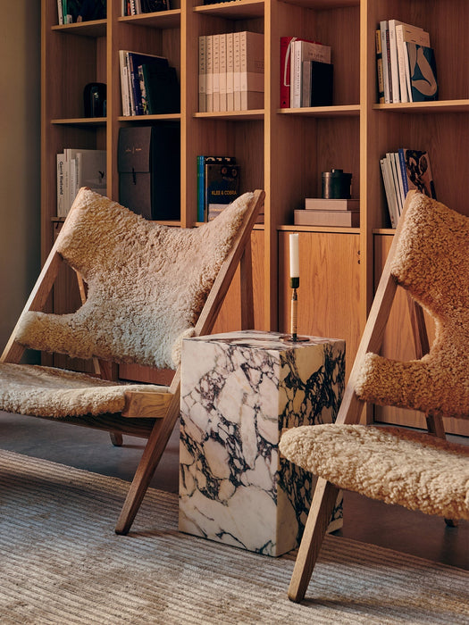 A living room with two Knitting Chairs, a marble Plinth table with a Duca Candle Holder, set on a Houkime Rug by a bookcase.