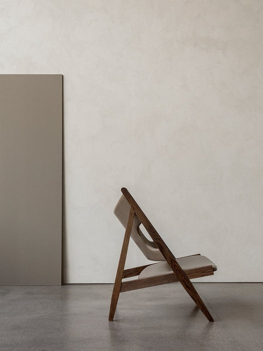 Side view of the Knitting Lounge Chair with a dark oak frame and light leather upholstery, against a textured off-white wall.