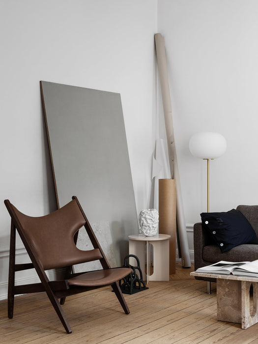 The Knitting Lounge Chair in brown leather and dark wood, styled in a minimalist room with a sofa, lamp, and side table.