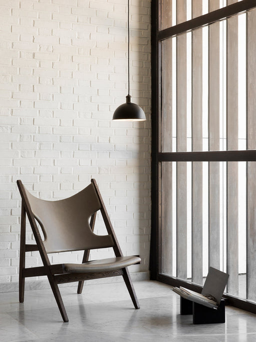 The Knitting Lounge Chair in dark wood and leather on a marble floor. A black Hubert Pendant hangs by a white brick wall.
