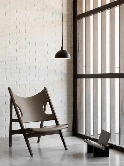 The Knitting Lounge Chair in dark wood and leather on a marble floor. A black Hubert Pendant hangs by a white brick wall.