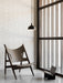 The Knitting Lounge Chair in dark wood and leather on a marble floor. A black Hubert Pendant hangs by a white brick wall.