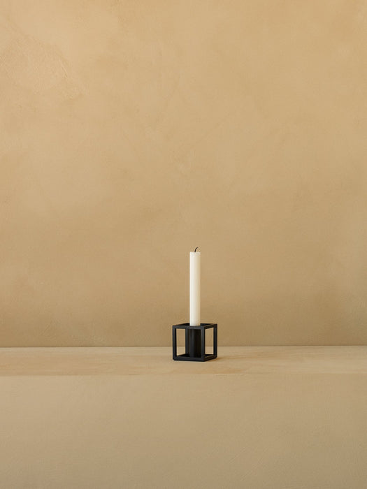 A single black Kubus T candleholder holds a white taper candle, sitting on a beige surface against a textured beige wall.