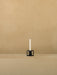 A single black Kubus T candleholder holds a white taper candle, sitting on a beige surface against a textured beige wall.