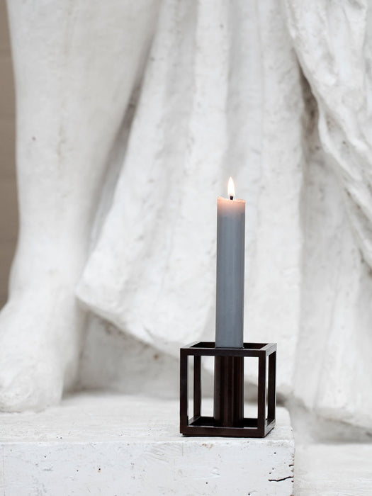 A lit grey by Lassen candle in a geometric, dark metal Kubus 1 candle holder on a white stone surface before a white statue.