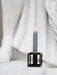 A lit grey by Lassen candle in a geometric, dark metal Kubus 1 candle holder on a white stone surface before a white statue.