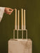 A hand with a match lights a cream candle in the off-white Kubus 4 Candle Holder, set upon a beige marble Plinth pedestal.
